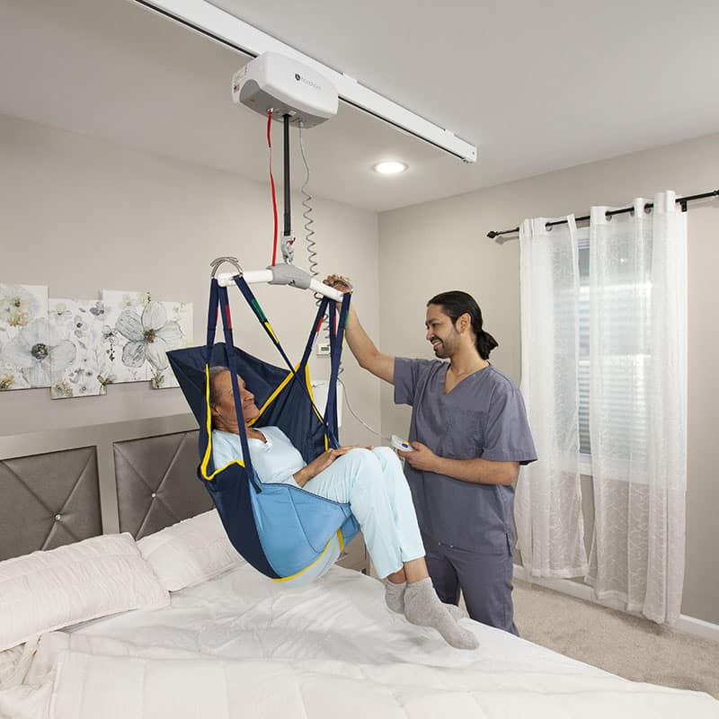 A nurse lowers an older woman down onto a bed using a patient lift