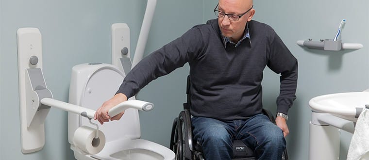 Man in a wheelchair rests his hand on a grab bar next to a toilet