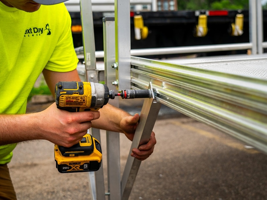 a person wearing a neon green shirt holds a yellow drill and drives a bolt into a metal beam