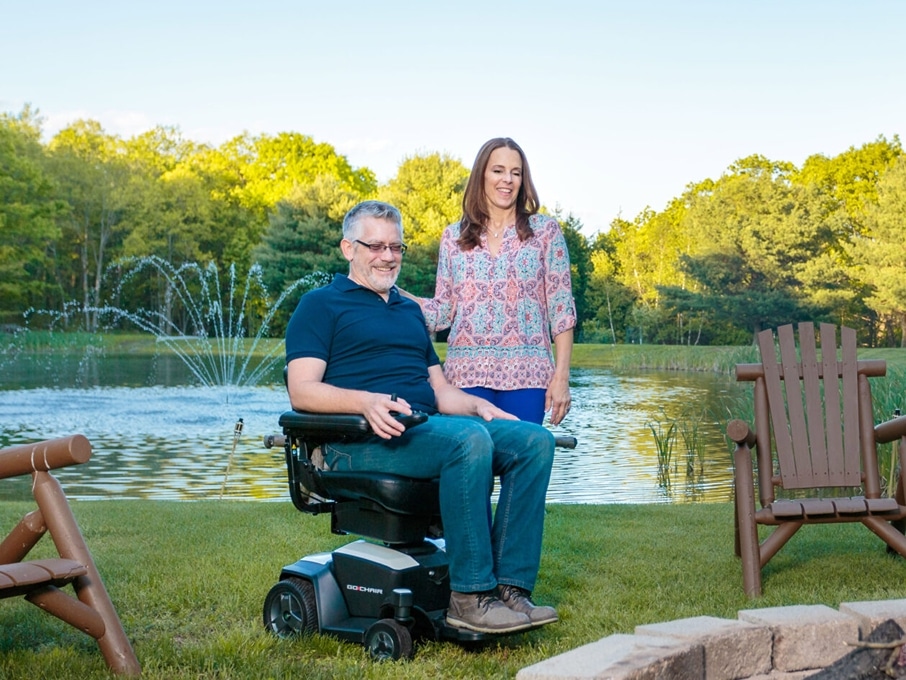 A man in a power wheelchair and his wife are outside in front of a bonfire with a fountain and pond in the background