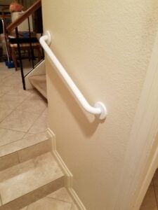 Curved white grab bar installed on a textured beige wall between a tile and a carpeted staircase.