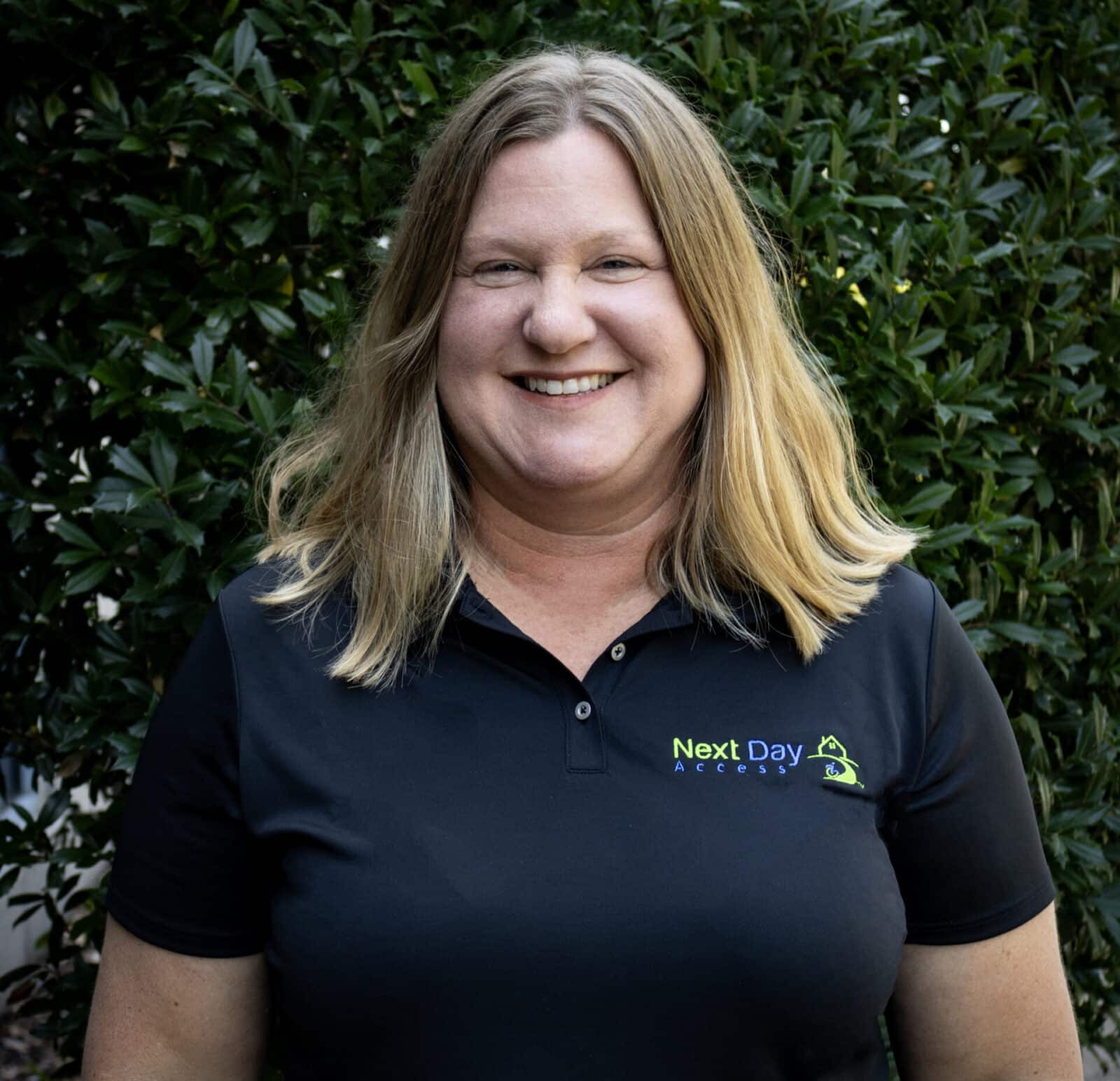 Professional headshot of a smiling woman with blonde hair, wearing a black polo shirt with the green and blue "Next Day Access" logo.