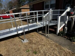 Elevated aluminum modular ramp extending from a white wooden porch railing of a brick home.