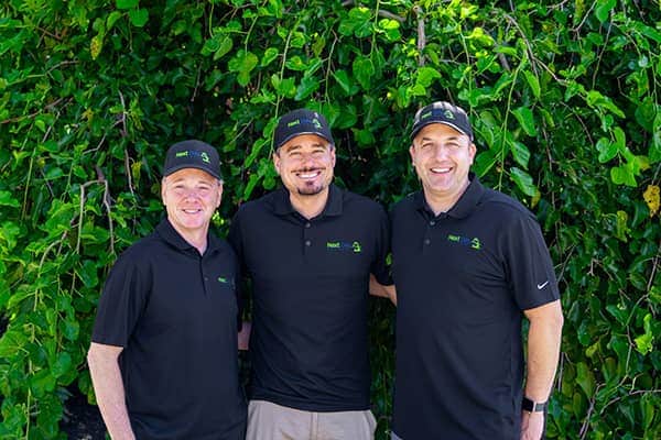 Three men, team members, wearing black Next Day Access logo polo shirts and hats, posing outdoors in front of green foliage.