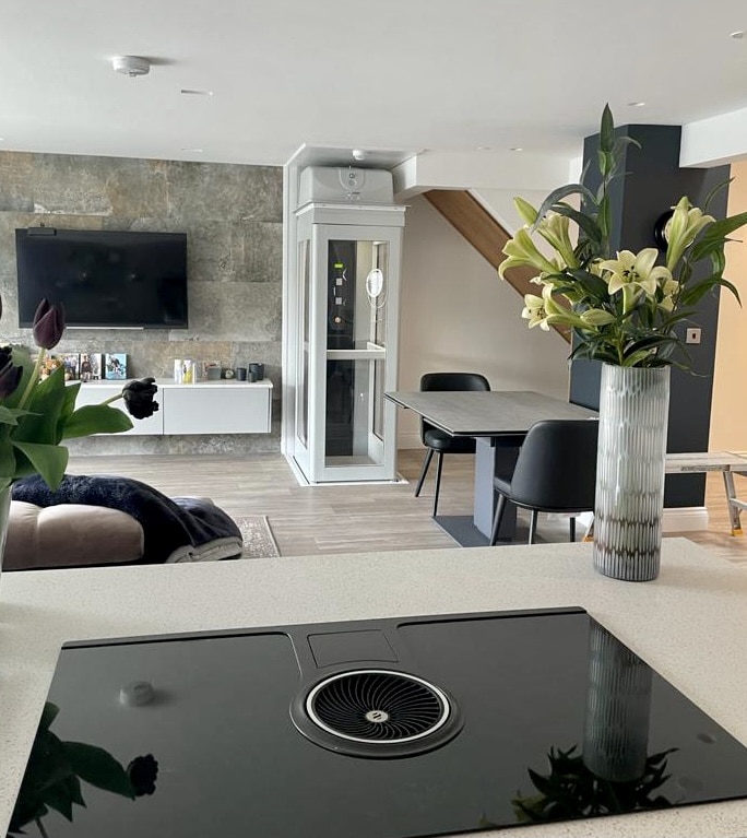 White and glass residential elevator cabin installed in an open-plan living room near stairs and a dining table.