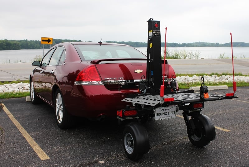 A vehicle lift on large rugged wheels being towed behind a red sedan