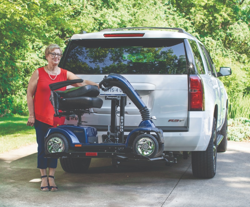 Woman in red stands next to a motorized scooter raised onto the back of her white SUV