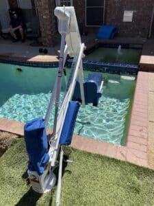 Portable white pool lift with blue padding and safety warning stickers by a rectangular brick-rimmed pool.