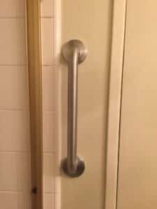 Vertical brushed stainless steel grab bar mounted on a beige wall next to a white tiled shower opening.