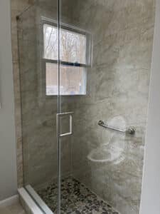 Frameless glass shower with neutral marble-look tile walls, a pebble floor, a window, and a silver grab bar.