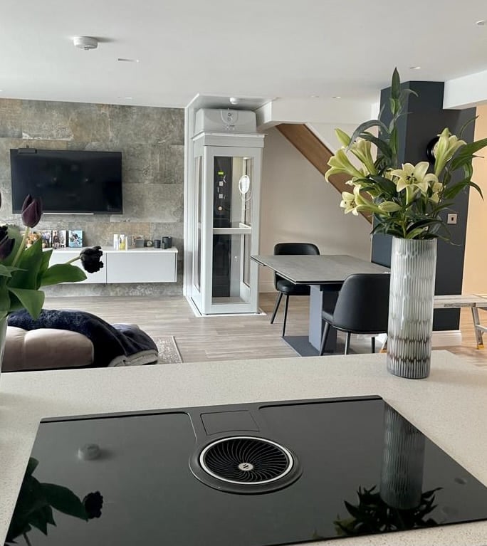 White and glass residential elevator cabin installed in an open-plan living room near stairs and a dining table.