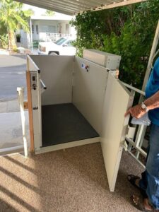 Open vertical platform lift with light beige metal walls and a dark floor, installed on a porch.