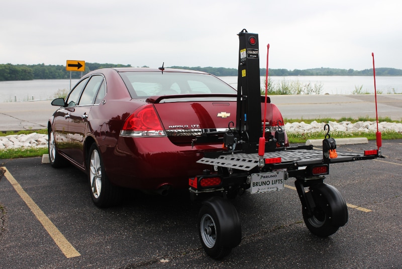 A vehicle lift on large rugged wheels being towed behind a red sedan