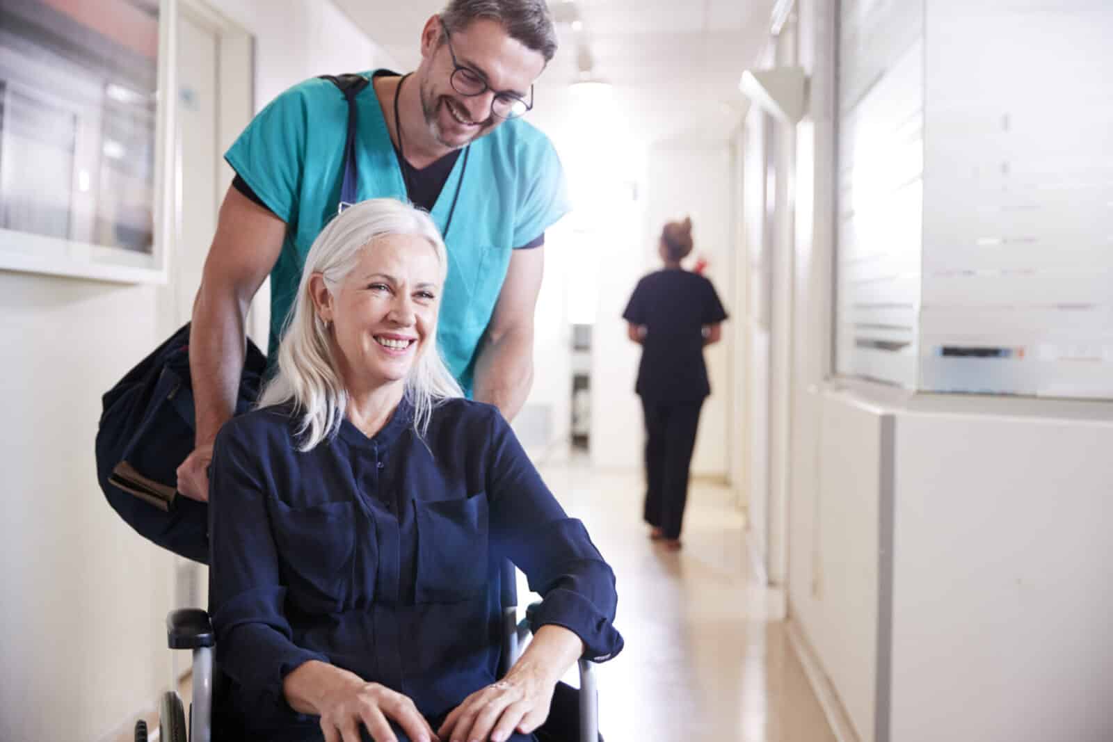 A smiling senior woman in a wheelchair being pushed by a male orderly in a hospital hallway.