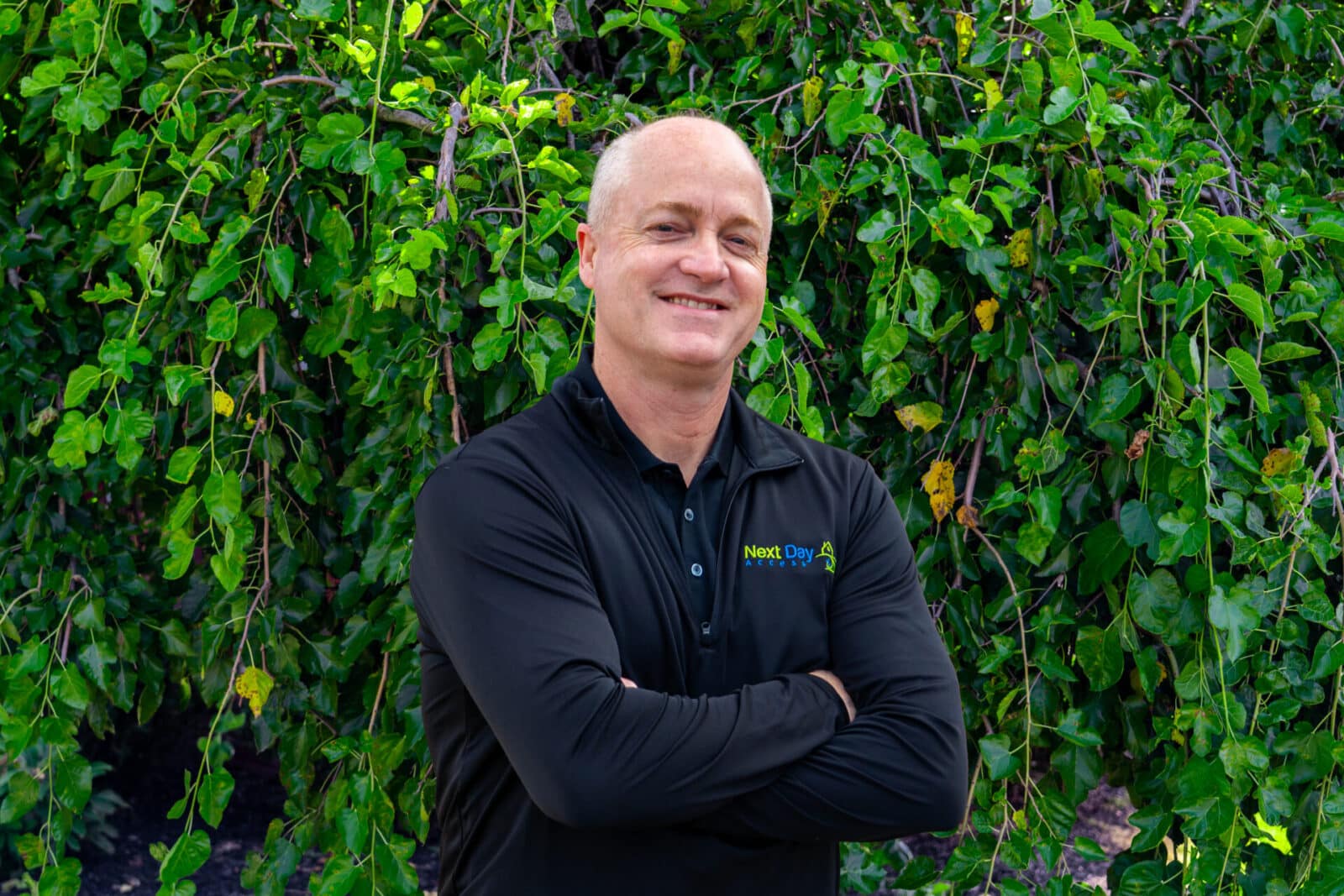 Smiling bald man with crossed arms in front of a leafy green tree, wearing a black shirt with the "Next Day Access" logo.