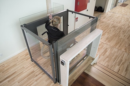 woman rides a glass wheelchair lift up to a large office space with a wooden floor