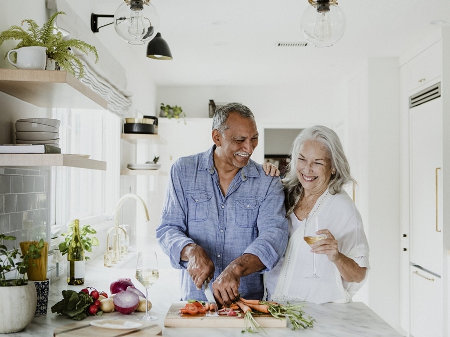 an elderly couple smile in the kitchen while cutting vegetables and drinking wine