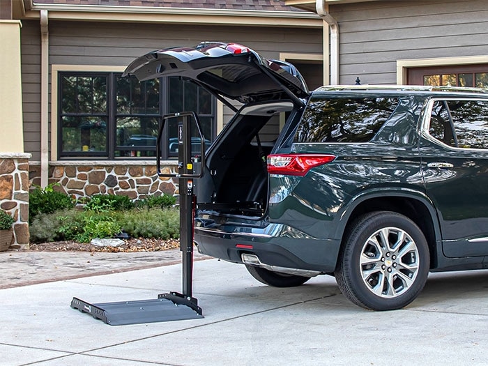 A lowered vehicle lift sits flat on the ground as it extends from the back of a mini van trunk