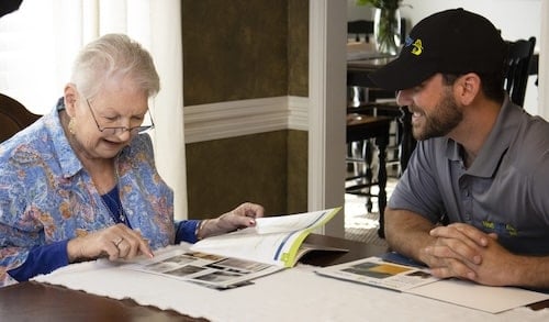 An older woman in a blue floral shirt and glasses looks at a magazine with a man in a black cap and gray shirt.