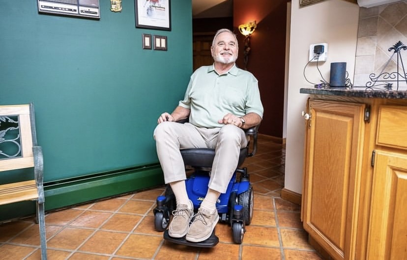 Man sits on a power wheelchair in his kitchen and faces the camera