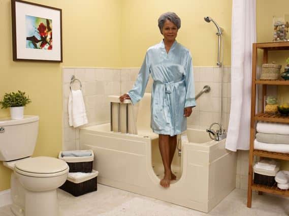 Woman in a bathrobe steps out of a walk-in tub in a bathroom with yellow walls