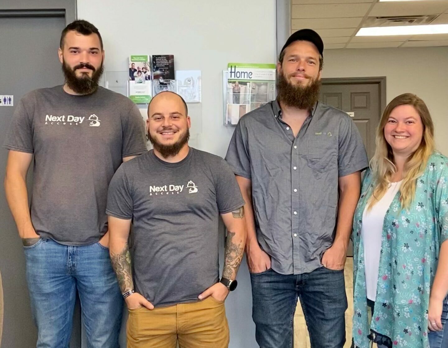 Four Next Day Access team members smiling and standing in front of an office wall with brochures.
