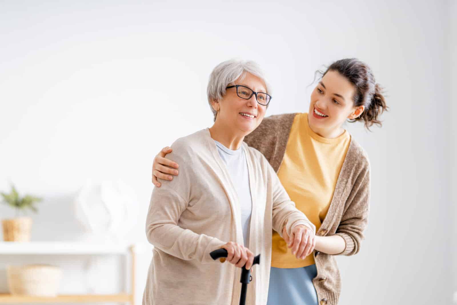 A younger woman gently embraces an older woman using a cane, conveying an impression of compassionate care.