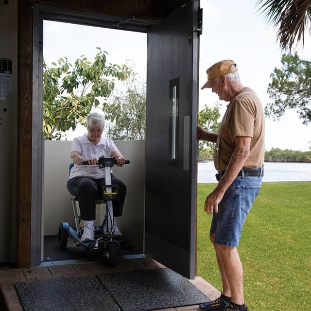 older man holds a wheelchair lift door open for a woman on a scooter