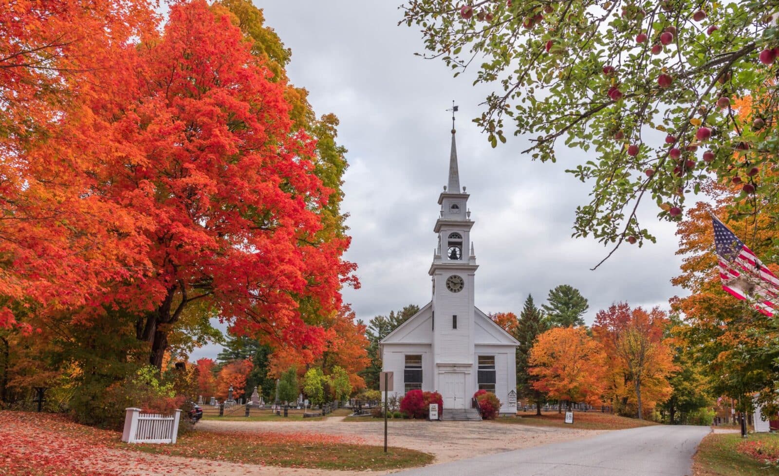 White church with a tall steeple and clock, surrounded by trees with brilliant red and orange autumn foliage in New Hampshire.