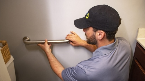 a Man in a uniform and baseball cap installs a handrail onto the bathroom wall
