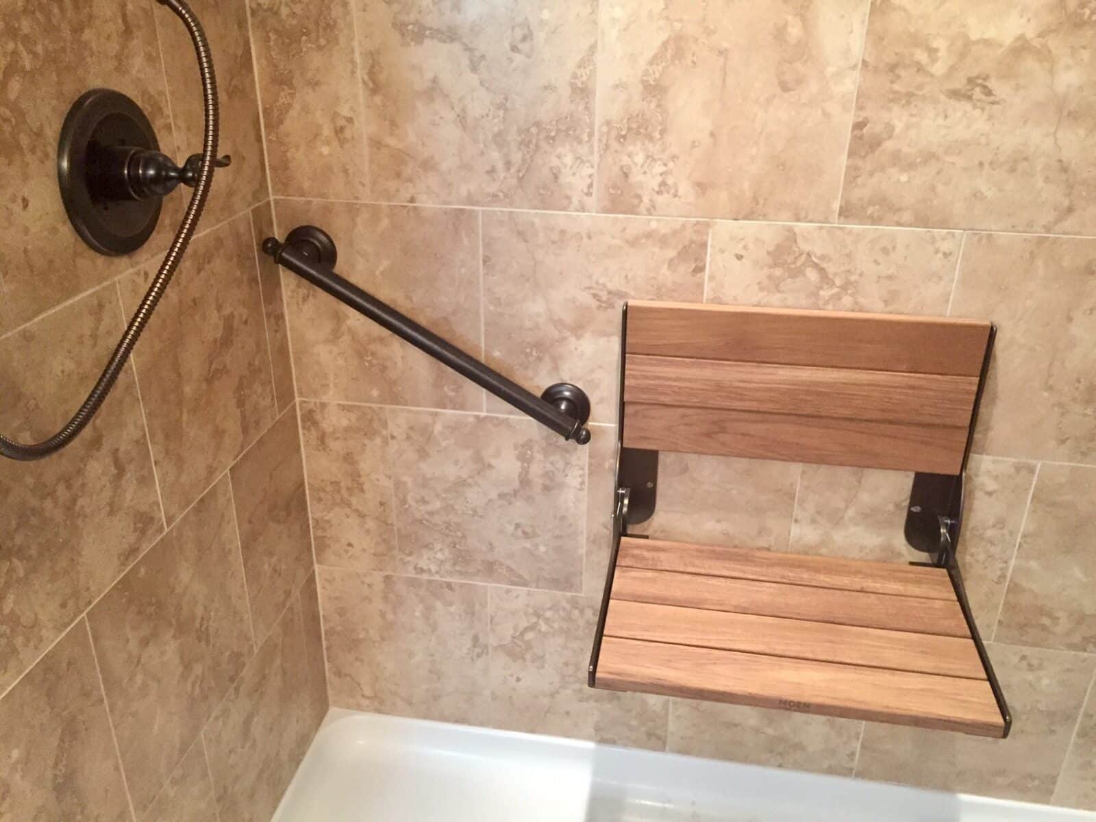 a folding wooden shower chair mounted to the wall of a tiled shower next to a grab bar