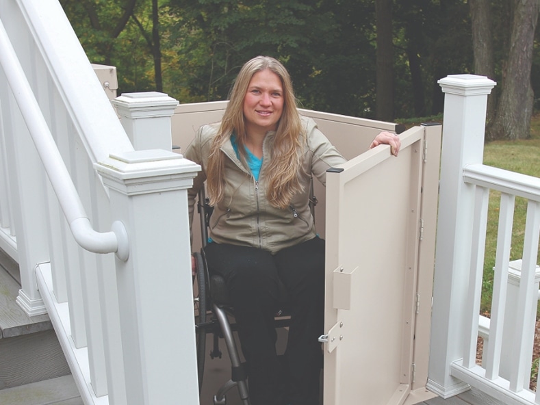 Woman in a wheelchair opens the door to her porch lift and exits onto her raised deck