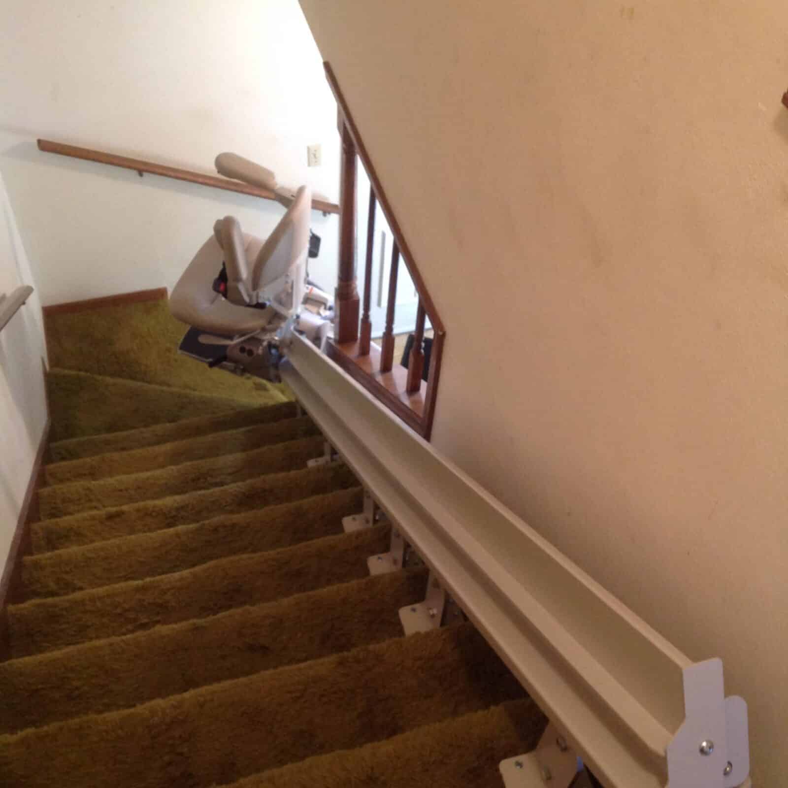 Beige curved rail stairlift on a patterned tile floor at the base of a shag carpeted staircase.