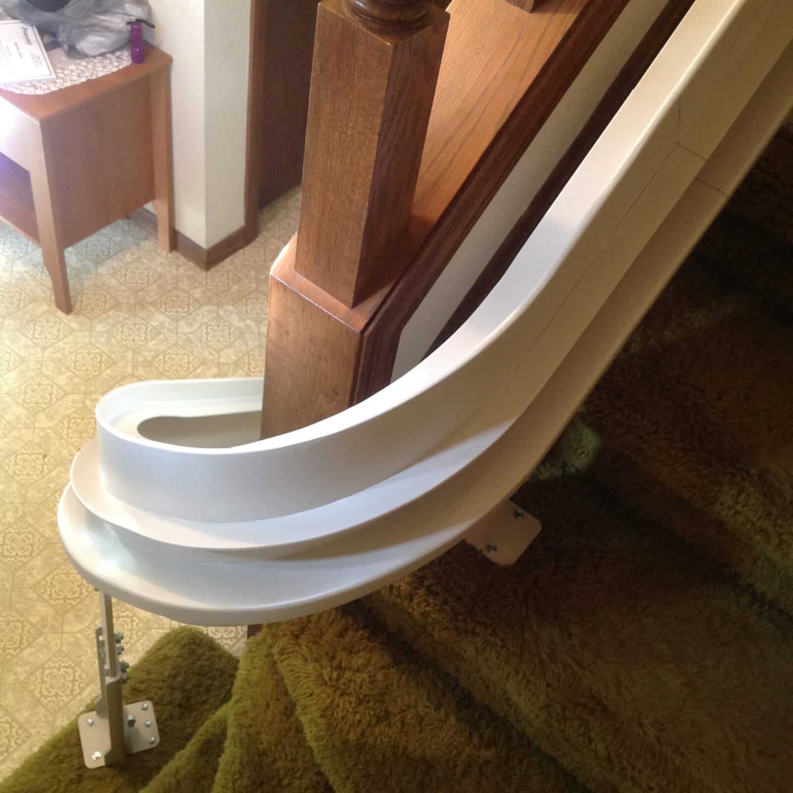 Beige curved rail stairlift on a patterned tile floor at the base of a shag carpeted staircase.