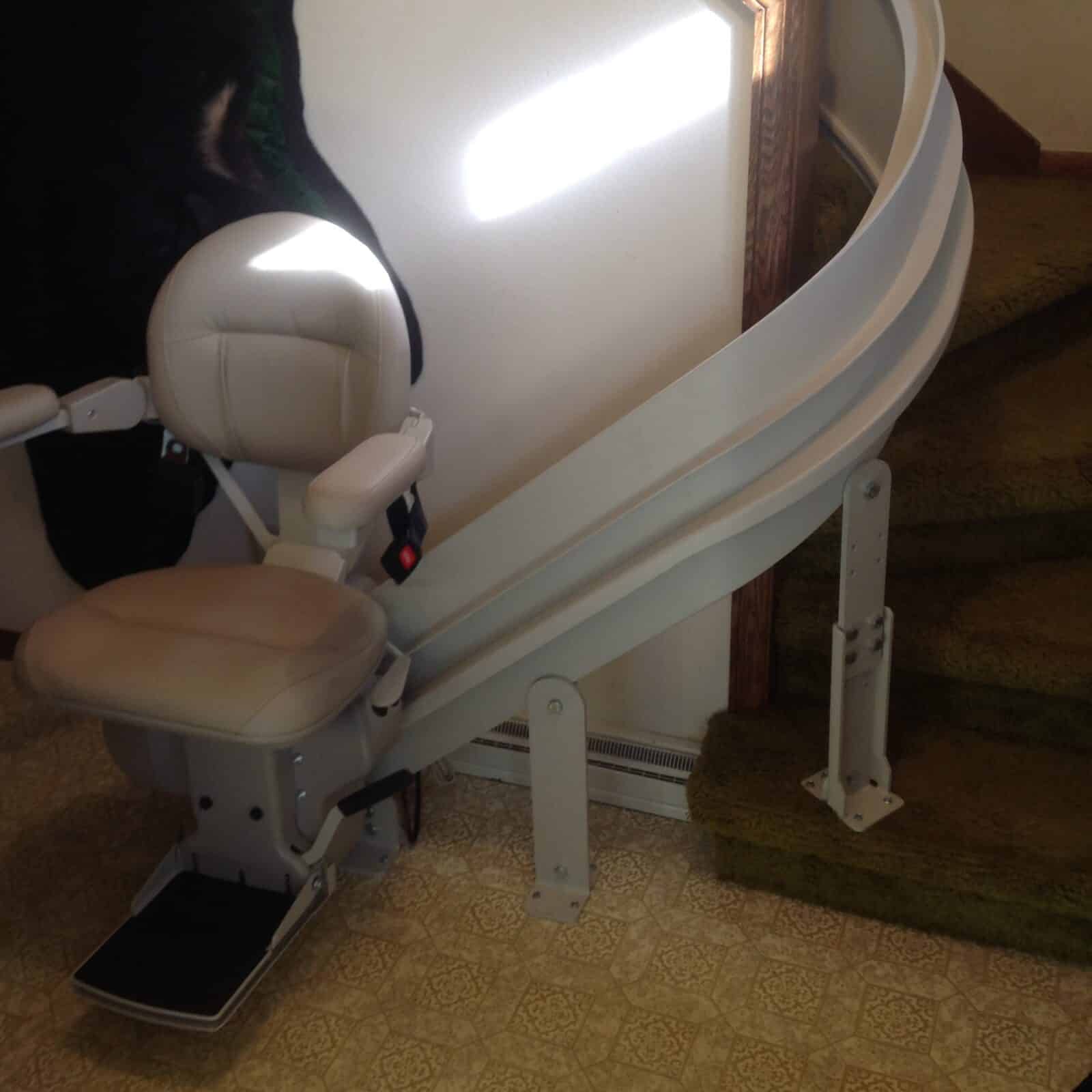 Beige curved rail stairlift on a patterned tile floor at the base of a shag carpeted staircase.