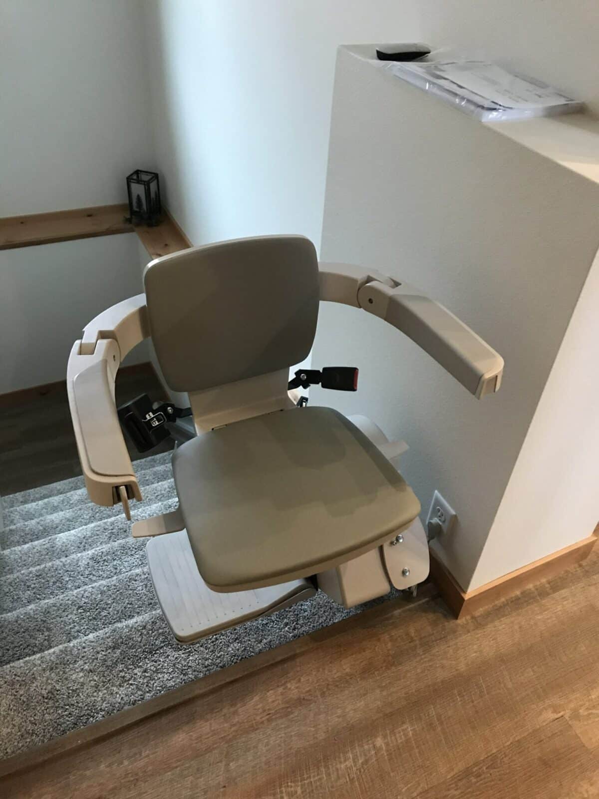Taupe stairlift chair with armrests and red safety buckle at the top of a gray carpeted staircase.