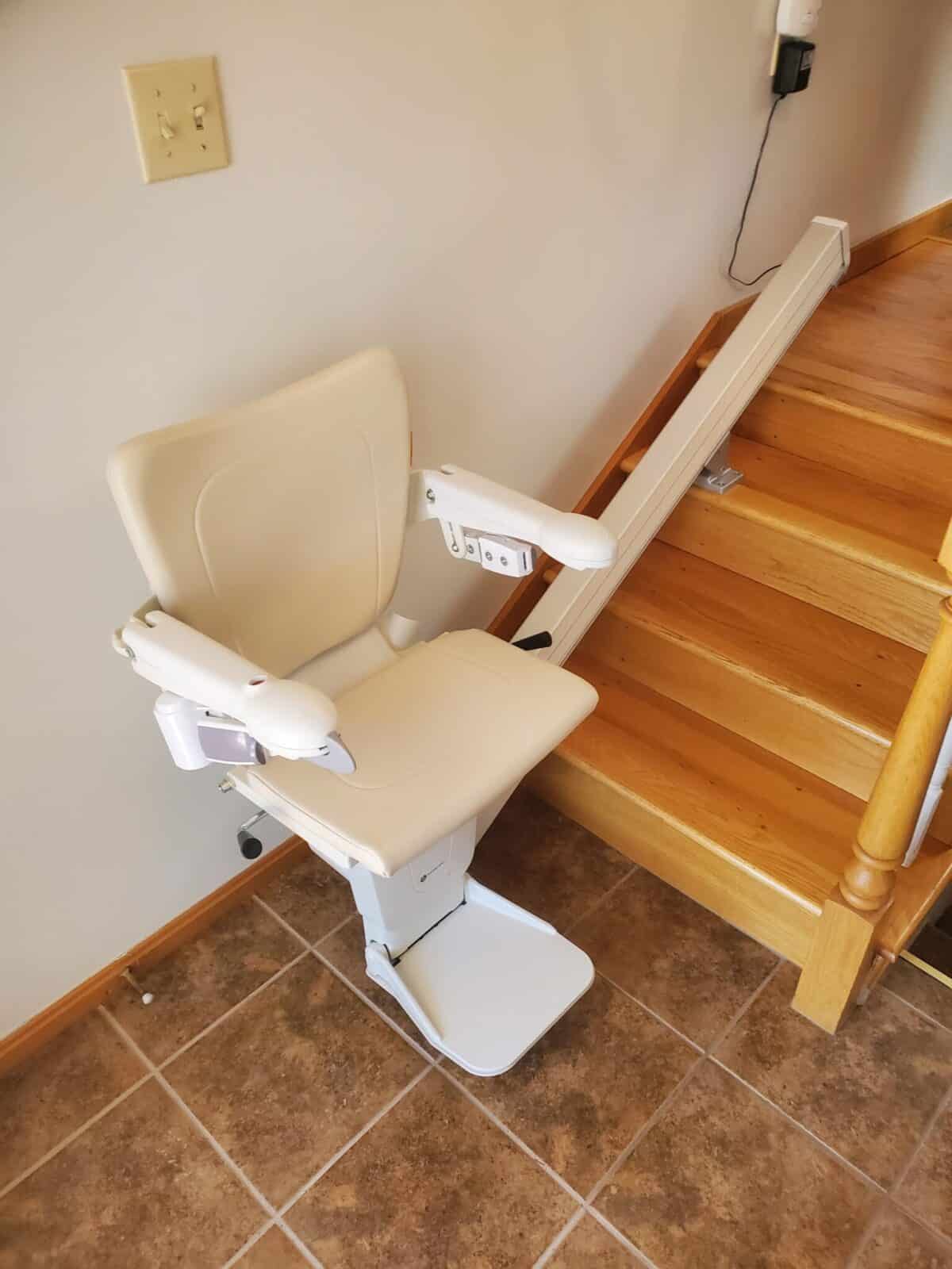 Light tan stairlift installed on straight white rail next to wooden stairs and a tiled landing.