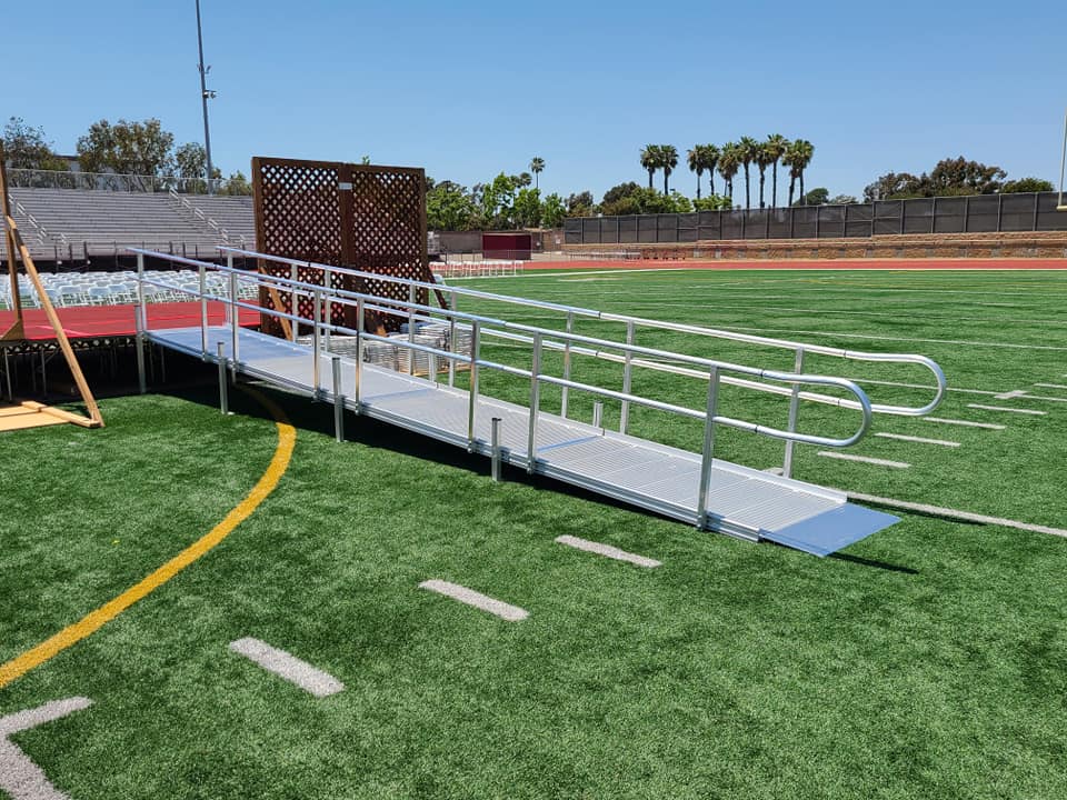 A modular aluminum wheelchair ramp with silver handrails extends onto a green artificial turf football field.