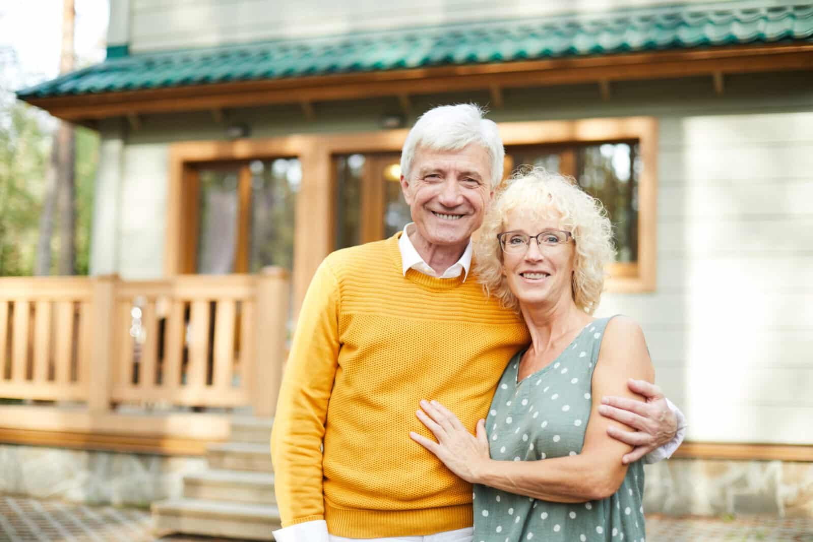 An older couple smiling in front of a rustic wooden house; the man wears a bright yellow sweater.