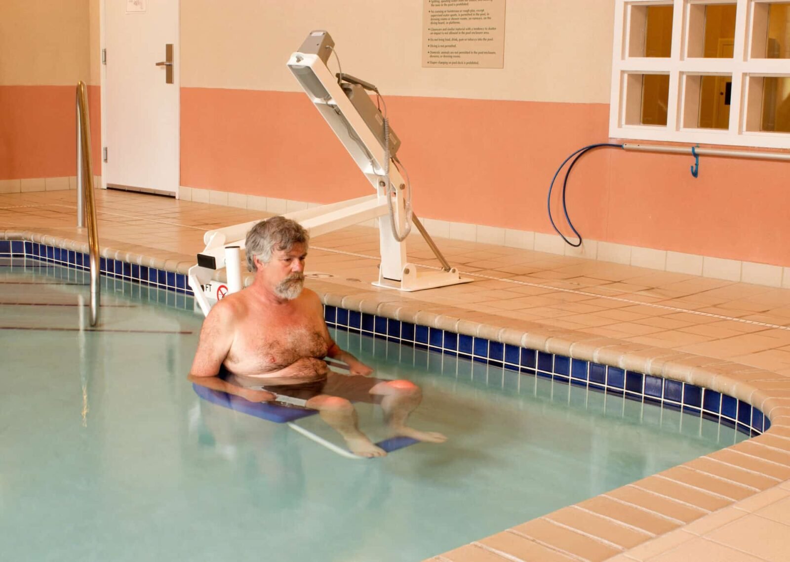 A blue pool lift installed next to a public hot tub with rules listed on the wall in the background