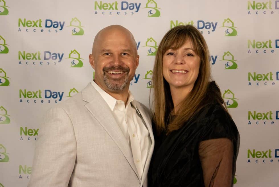Bald man in light suit and woman in black smile in front of a white backdrop with repeating "Next Day Access" logos.