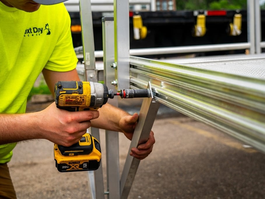 A worker in a yellow 'Next Day Access' shirt uses a power drill to assemble a ridged aluminum ramp.