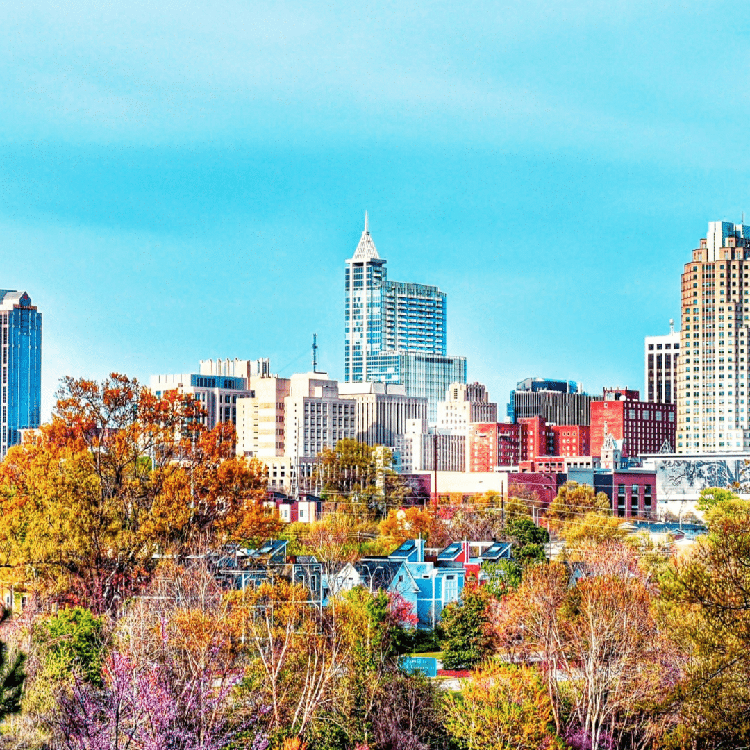 The Raleigh-Durham skyline with tall buildings rising above dense, brightly colored autumn foliage.