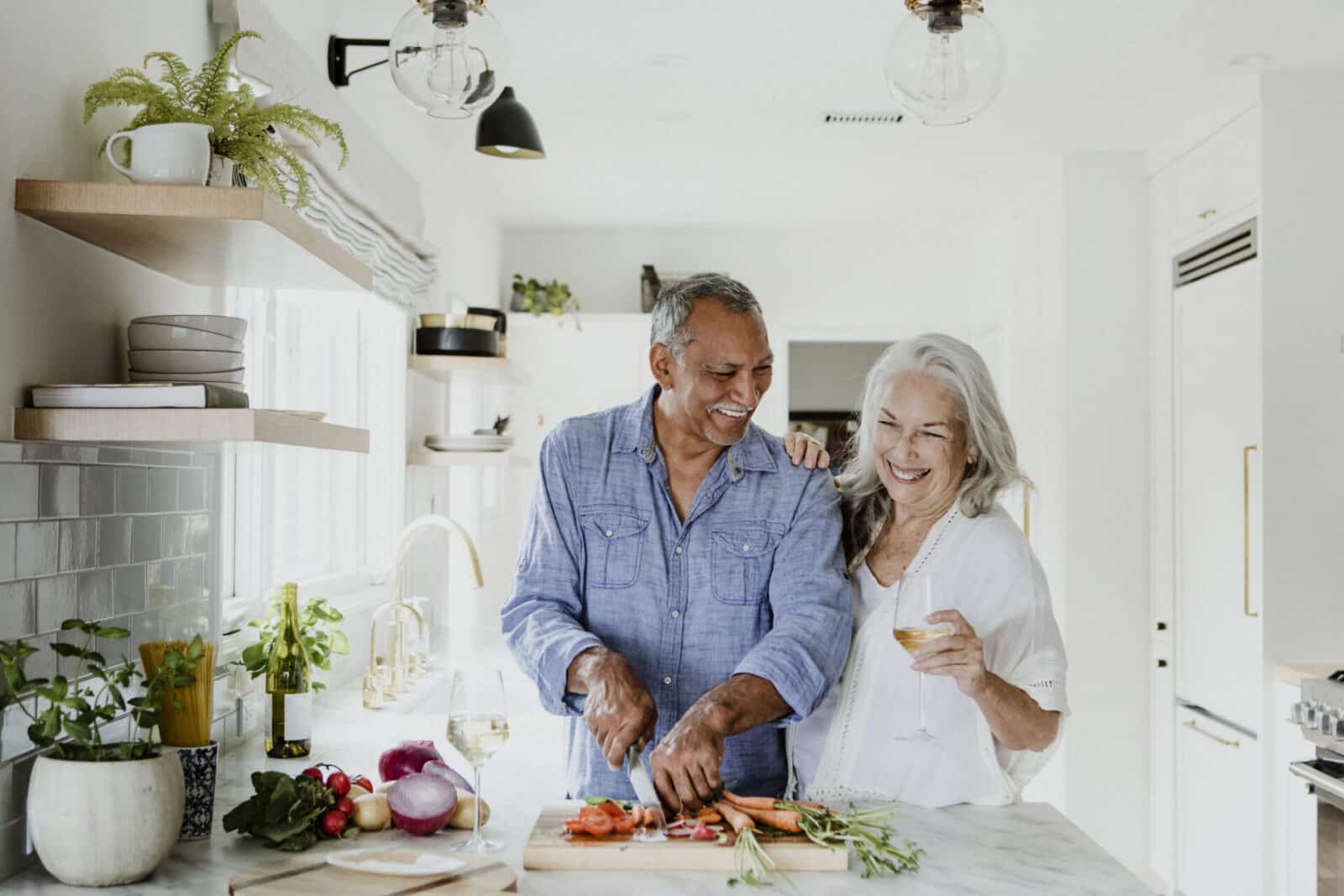 Elderly couple cooking in a bright kitchen; the man chops vegetables while the woman holds a wine glass.