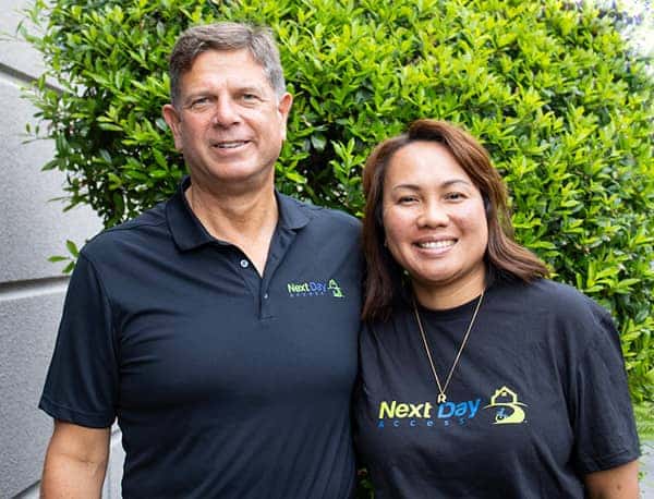 Man and woman in black "Next Day Access" shirts smiling outdoors in front of a green hedge.