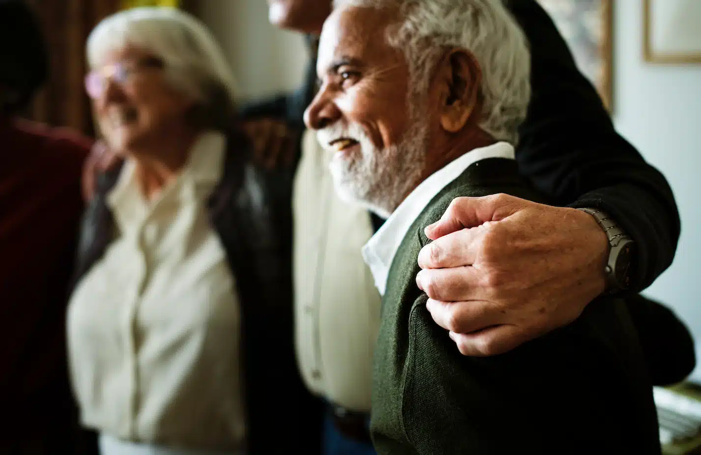 A smiling, bearded older man in a green sweater with an arm resting on his shoulder