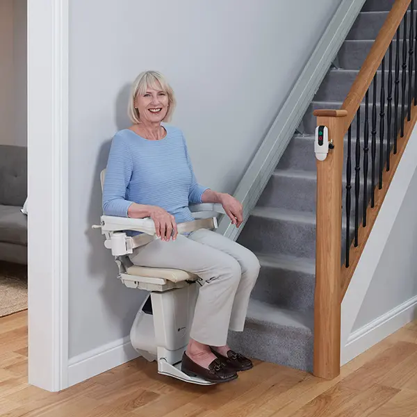 1100-straight-stairlift-woman-in-use-1.jpg Woman in a blue sweater smiles and sits at the bottom of the stairs on a chair lift