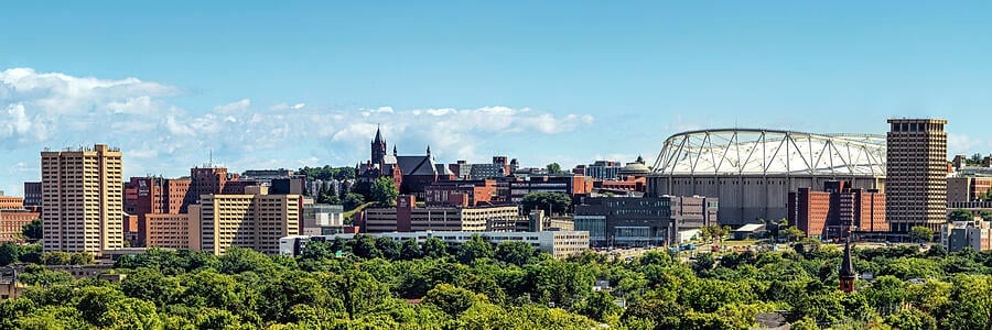 Panoramic daytime view of the Syracuse skyline featuring university buildings, lush green trees, and the JMA Wireless Dome.