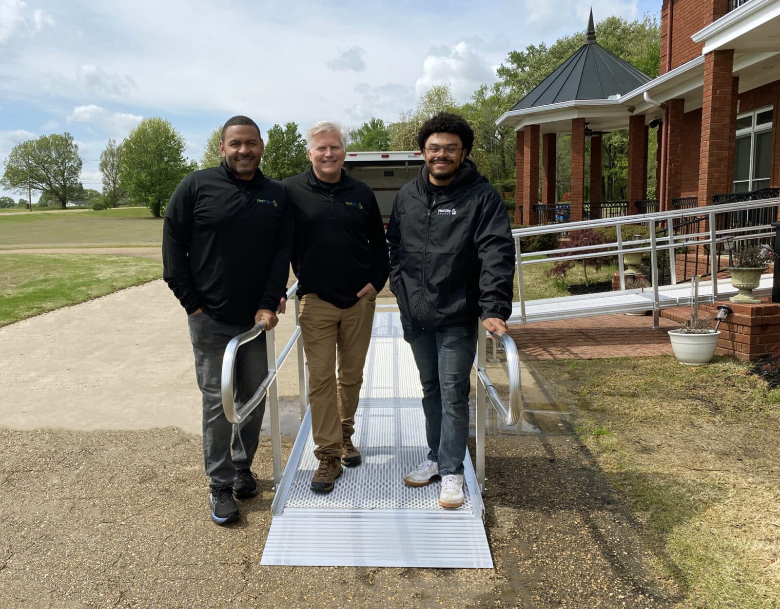 Three men in Next Day Access apparel standing on a small aluminum access ramp system outside a red brick building with a gazebo.