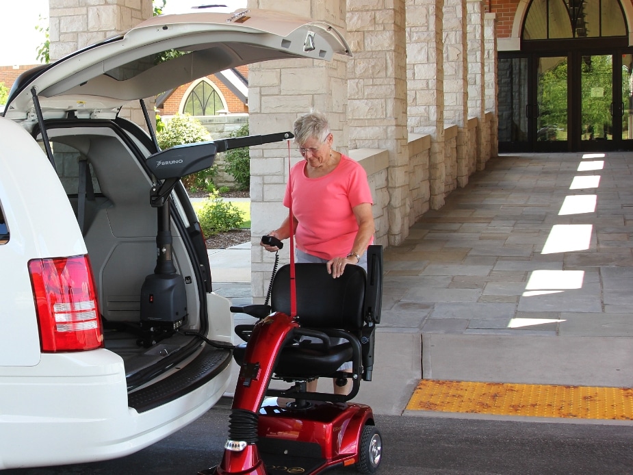 A woman uses a hoist to lift a power scooter into the back of her white van.
